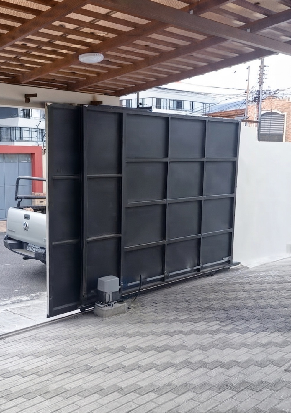 Port&oacute;n corredizo negro de paneles met&aacute;licos, con motor de cremallera a la vista, bajo p&eacute;rgola de madera